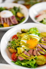 A fresh gourmet salad with sliced beef, cherry tomatoes, potatoes, leafy greens, and a poached egg drizzled with green dressing, served in a bowl. Other salads are blurred in the background