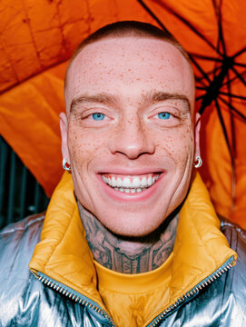 Bright smile of young man with freckles, blue eyes, and shaved head, wearing shiny silver jacket under orange umbrella. His joyful radiates positivity