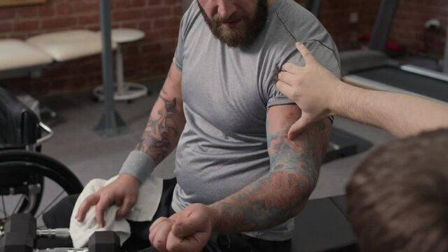 Unrecognizable personal trainer examining shoulder muscle of bearded bodybuilder with disability while they having workout