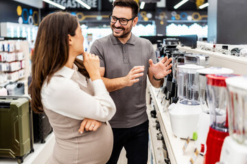 Smiling shop assistant helping a pregnant customer choose home appliances while showcasing various products in a vibrant electronics store
