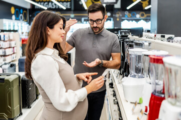Smiling shop assistant helping a pregnant customer choose home appliances while showcasing various products in a vibrant electronics store