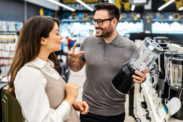 Smiling shop assistant helping a pregnant customer choose home appliances while showcasing various products in a vibrant electronics store