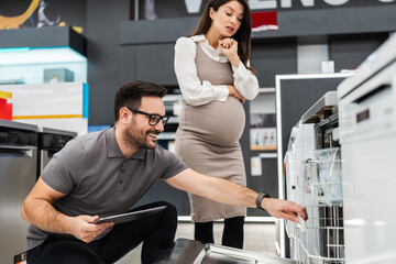 Smiling salesman holding a tablet, discussing and showing dishwashers to a pregnant customer, selecting the best options in a modern home appliance store