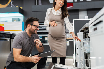Smiling salesman holding a tablet, discussing and showing dishwashers to a pregnant customer, selecting the best options in a modern home appliance store