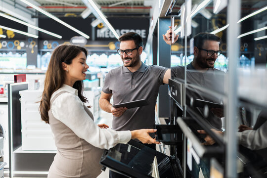 Friendly salesman holding a digital tablet is showing different modern ovens to a pregnant customer in a home appliances store, they are both smiling and looking at the oven