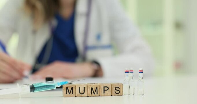 Letters MUMPS on wooden blocks with vials and syringe on doctor workplace. Female healthcare worker prescribes vaccine sitting at hospital desk