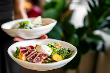 A person holds two bowls of fresh gourmet food. The front bowl contains seared tuna, a poached egg, greens, cherry tomatoes, and roasted potatoes. The background bowl features another dish