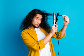 Man in yellow shirt styling long brunet hair using a flat iron against a bright blue background