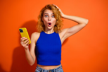 Obraz premium Young woman in a blue top displaying surprise while holding a yellow smartphone against a vibrant orange background