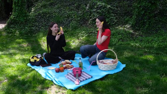 Young women pretend phone call with bananas for joke enjoying picnic in park. Happy female friends have fun together eating out on blue blanket