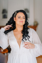Naklejka premium Confident bride in a white gown touching her hair, with styled curls and a delicate necklace, radiating elegance on her wedding day indoors..
