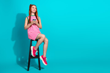 Cheerful young woman sitting on stool against blue background wearing pink checkered dress and red sneakers holding drink
