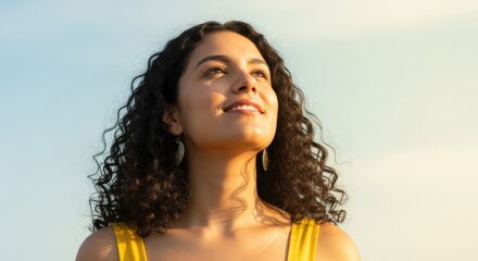 Close-up portrait of a woman looking up with joy against a clear sky background with sun glare, wearing a yellow top, and natural lighting.