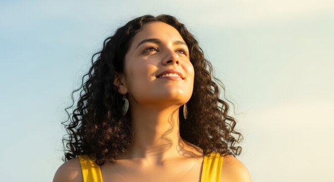 Close-up portrait of a woman looking up with joy against a clear sky background with sun glare, wearing a yellow top, and natural lighting.