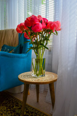 Bright pink peonies in a clear glass vase on a rustic wooden table, cozy interior with teal armchair, houseplants, and soft natural light. Homey and vibrant atmosphere.