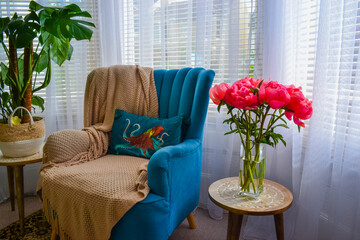 Bright pink peonies in a clear glass vase on a rustic wooden table, cozy interior with teal armchair, houseplants, and soft natural light. Homey and vibrant atmosphere.