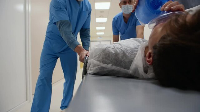 Tracking shot of medical staff in scrubs pushing patient on gurney while administering oxygen, moving urgently through hospital corridor during critical care situation