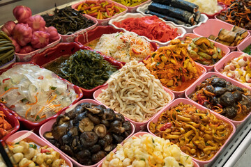 Bright assortment of various pickled vegetables, mushrooms, and seaweed displayed in colorful containers at a bustling food market stall.