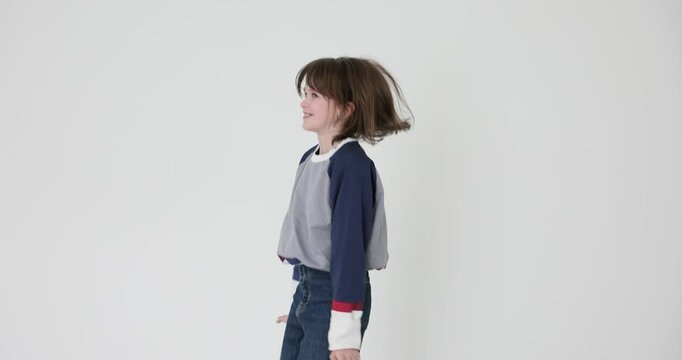 Cheerful little girl with messy hair jumps turning around at white wall. Playful schoolgirl in colorful sweatshirt and jeans has fun slow motion