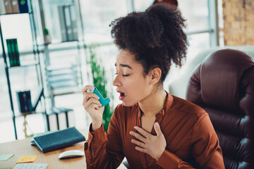 Young businesswoman using an inhaler in a modern office workspace with natural light, highlighting professional attire
