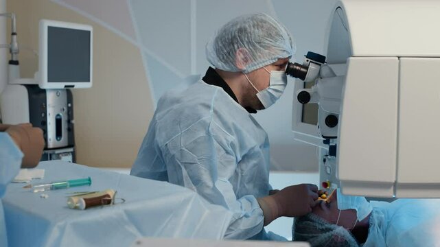 Surgeon using high-precision microscope to perform laser eye surgery on lying patient, while his assistant providing tools in sterile ophthalmic operating room