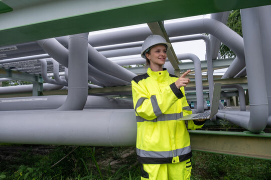 Engineer in safety gear checks data on laptop beneath industrial piping system at a construction site during daytime