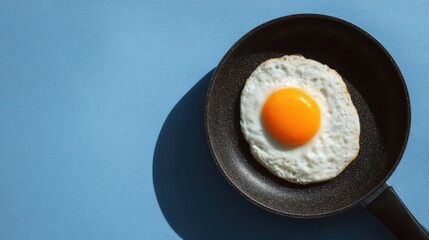 A perfectly cooked fried egg sits in a black frying pan against a light blue background. The bright yellow yolk stands out, showcasing a classic breakfast dish ready to serve