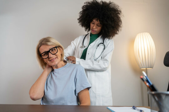 A mature Caucasian woman is having her neck examined by a Black female doctor in a clinic, due to stiffness, muscle spasm, or similar neck issues during a medical checkup.