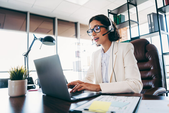 Confident businesswoman in formal attire working at her office desk with a headset and laptop, managing tasks.