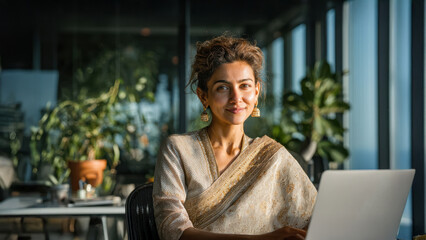 An 30s Indian woman wearing elegant traditional attire sitting confidently at a sleek modern office desk, working on a laptop.