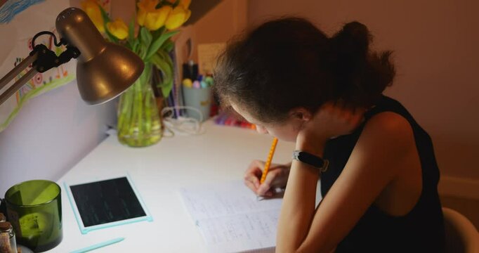 Female student sits at desk studying hard and writing in quiet evening. Paper notebook with tablet lie in front of girl while lamp casts warm glow on workspace
