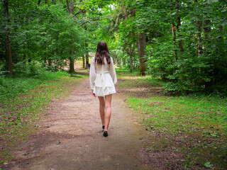 Stylish girl is walking down a path in a forest park. She is wearing a white dress and black shoes....