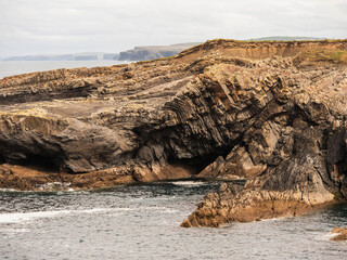 A rocky cliff coastline by the ocean. The water is calm and the sky is overcast. The scene is...
