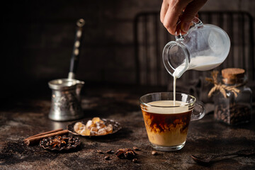 Coffee in a glass with cream poured over, sugar and coffee beans on dark background