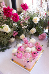 Rectangular pink frosted cake decorated with fresh pink and white ranunculus flowers and edible blossoms on white table. Studio food photography. Wedding dessert and floral styling concept. Design for
