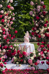 Two-tier pink and white wedding cake on white round table surrounded by red, white, and pink roses and hydrangeas in outdoor floral arch setup. Garden event photography. Wedding ceremony and celebrati