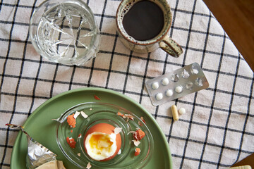 Top view of a breakfast on the table with a boiled egg, coffee, a glass of water, and pills