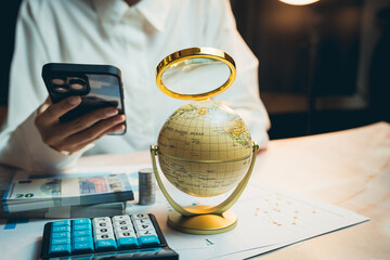 Business concept. Person Using Smartphone with Globe, Calculator, Cash, and Charts on Desk in...