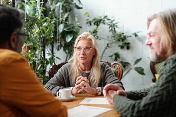 Senior blonde woman engaging in discussion with two colleagues over coffee in a bright office with...