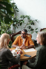 Group of three people sitting at round table, discussing plans in cozy indoor garden setting, surrounded by lush green plants, look relaxed and engaged in conversation