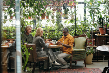 Seniors of diverse backgrounds sitting in a plant-filled cafe, engaging in conversation, and...