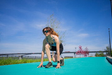 Concentrating female athlete preparing for a run on a vibrant green track in a bustling city park, surrounded by a clear blue sky, showcasing fitness and unwavering determination