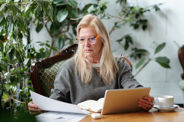 Senior Caucasian woman studying documents while holding a tablet in cozy room filled with plants and natural elements