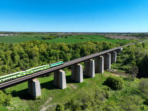 Commuter train travelling across a railway trestle bridge above Credit River, surrounded by lush forests and farmland in rural Ontario, Canada on bright spring day, blue sky, and scenic countryside