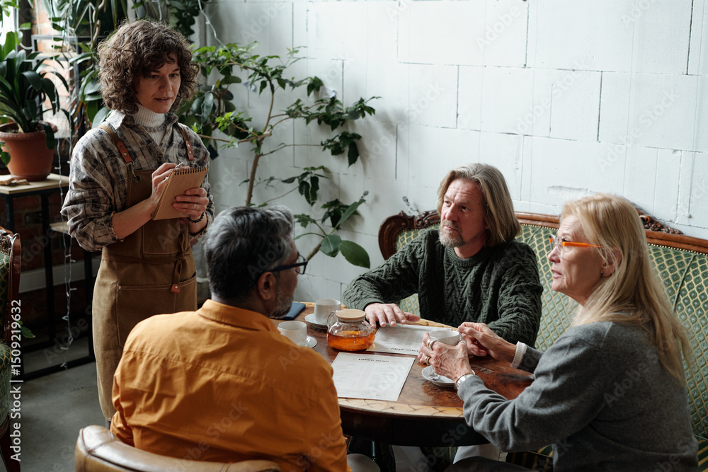 Poster group of colleagues engaging in discussion around wooden table in cozy office setting with plants an