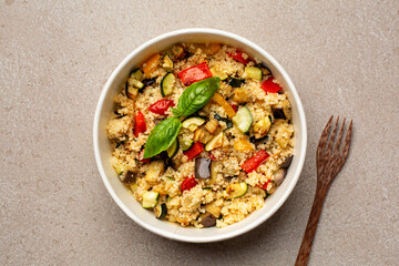 Couscous with roasted vegetables,  zucchini, egg plant, yellow and red bell pepper, basil. Round lunchbox with healthy food, prep meal. Top view, beige table.