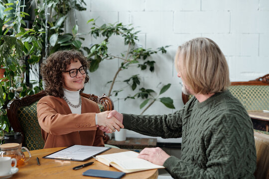 Two professionals shaking hands across a table in cozy, casual business setting with plants in background. Both have documents and notebooks on table, creating friendly and productive aura