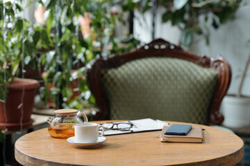 Scene showing a cozy corner with comfortable chair, pot of tea, cup, and open book on wooden table, surrounded by potted plants and natural greenery
