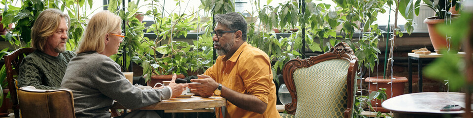 Three people sitting in a lush indoor cafe engaged in serious discussion among potted plants and...