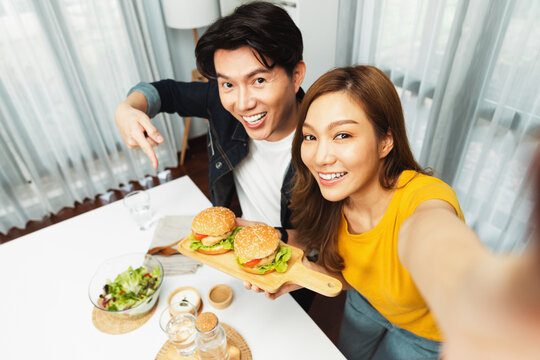 Top view of smiling host chef influencers taking photo with smartphone by selfie holding double hamburger on cooking show special easy with salad side dish homemade on social media channel. Infobahn.